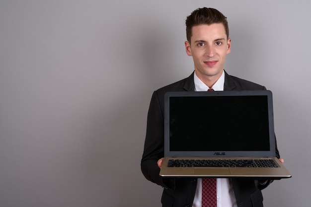 junger hübscher Geschäftsmann, der Anzug trägt, während Laptop auf Grau hält