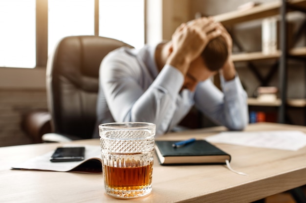 Junger hübscher Geschäftsmann sitzt am Tisch und leidet unter Kater in seinem eigenen Büro. Er hält die Hände auf dem Kopf. Vorne steht ein Glas Whisky.