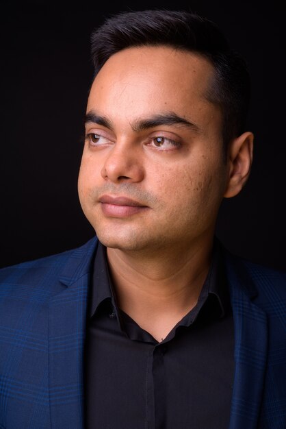 junger hübscher indischer Geschäftsmann, der blauen Anzug gegen schwarze Wand trägt