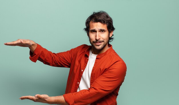 Junger hübscher indischer Mann, der ein Objekt mit beiden Händen auf Seitenkopierraum hält, ein Objekt zeigend, anbietet oder annonciert