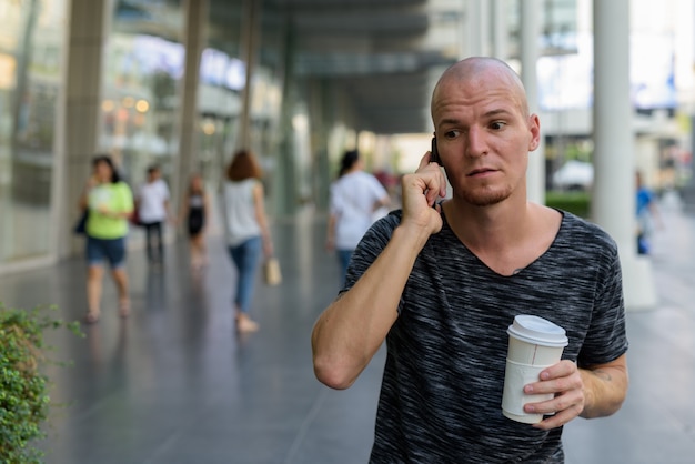 Junger hübscher kahler Mann, der Papierkaffeetasse hält, während er am Telefon spricht