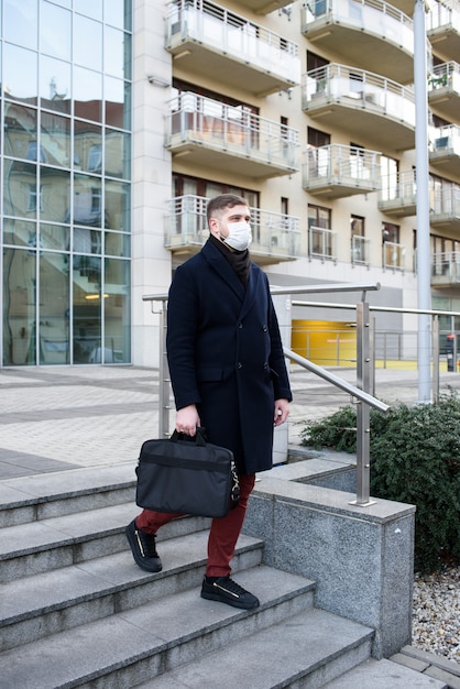 Junger hübscher kaukasischer Mann nahe seinem Büro, der chirurgische Gesichtsmaske trägt, um vor Coronavirus zu schützen