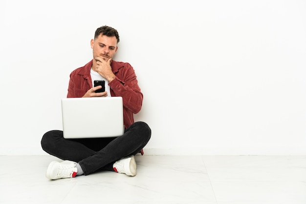 Junger hübscher kaukasischer Mann sitzt auf dem Boden mit Laptop, der denkt und eine Nachricht sendet