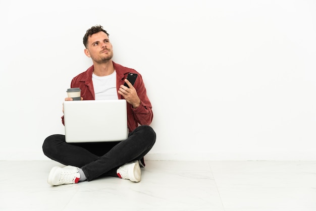 Junger hübscher kaukasischer Mann sitzt auf dem Boden mit Laptop, der Kaffee hält