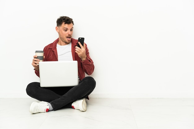 Junger hübscher kaukasischer Mann sitzt auf dem Boden mit Laptop, der Kaffee hält