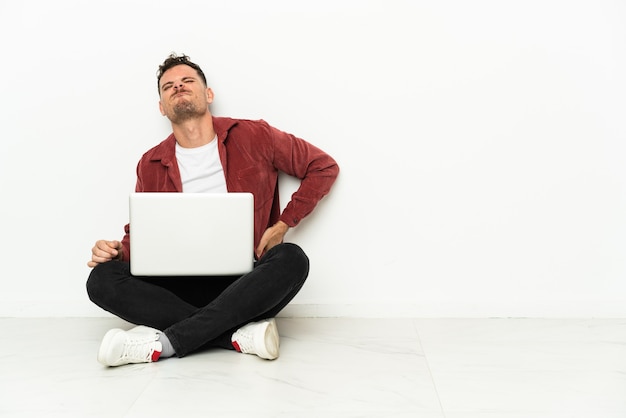 Junger hübscher kaukasischer Mann sitzt auf dem Boden mit Laptop, der unter Rückenschmerzen leidet, weil er sich bemüht hat