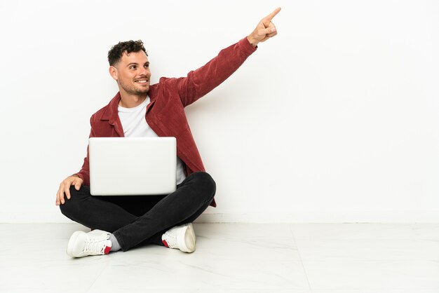 Junger hübscher kaukasischer Mann sitzt auf dem Boden mit Laptop, der weg zeigt