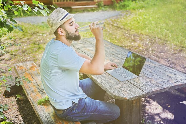 Junger hübscher kaukasischer Mann sitzt den Holztisch im Wald und trinkt Wasser, während er am Computer arbeitet.