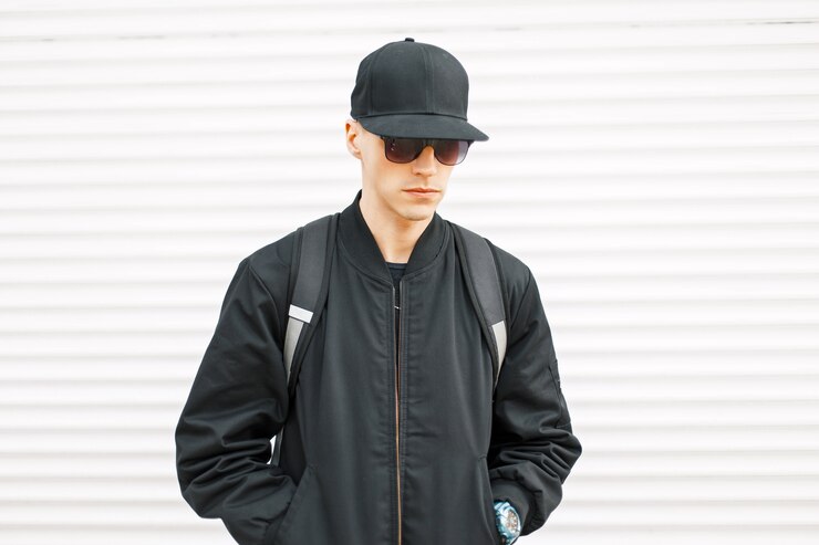Foto junger hübscher kerl in einer schwarzen jacke mit einer baseballkappe und einem rucksack nahe einer weißen metallwand