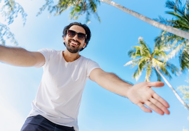 Junger hübscher lateinamerikanischer Mann in einem weißen T-Shirt und einer Sonnenbrille auf blauem Himmel und Palmen, streckt seine Hand zur Linse aus, Bild von unten