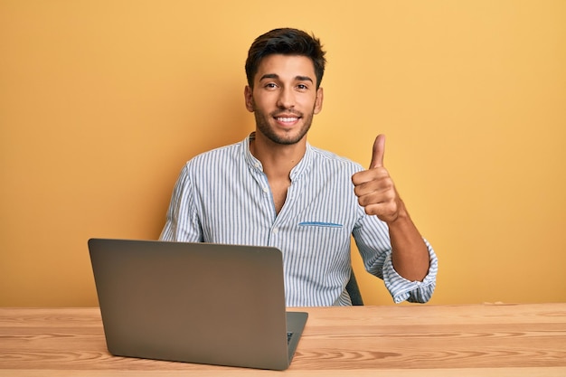 Junger hübscher Mann arbeitet im Büro mit Laptop lächelnd glücklich und positiv Daumen nach oben macht ausgezeichnet und Genehmigungszeichen