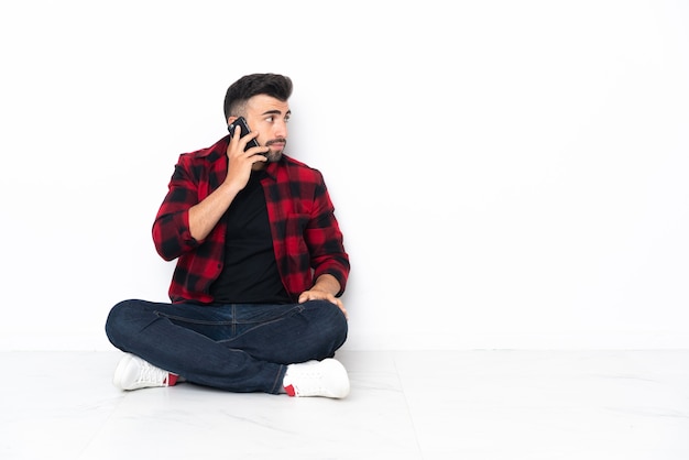 Junger hübscher Mann, der auf dem Boden sitzt und ein Gespräch mit dem Mobiltelefon mit jemandem hält