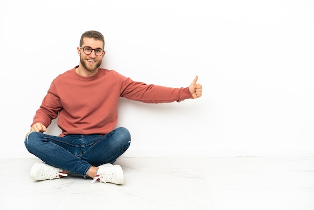 Junger hübscher Mann, der auf dem Boden sitzt und eine Daumen hoch Geste gibt