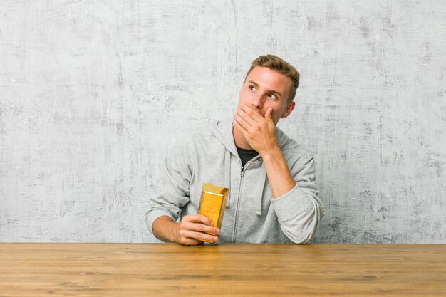 Junger hübscher Mann, der einen Goldbarren auf einem Tisch hält, der nachdenklich zu einem leeren Raum schaut, der Mund mit Hand bedeckt.