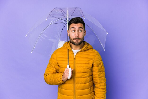 Junger hübscher Mann, der einen Regenschirm isoliert hält, verwirrt, fühlt sich zweifelhaft und unsicher.