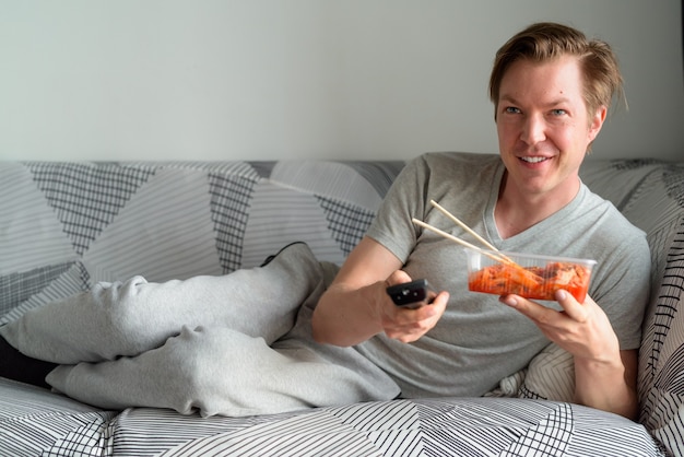 Junger hübscher Mann, der fernsieht und Kimchi im Wohnzimmer zu Hause isst