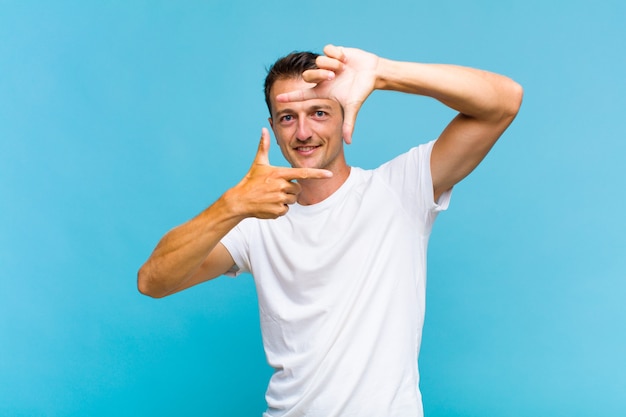 Junger hübscher Mann, der glücklich, freundlich und positiv fühlt, lächelt und ein Porträt oder Fotorahmen mit Händen macht