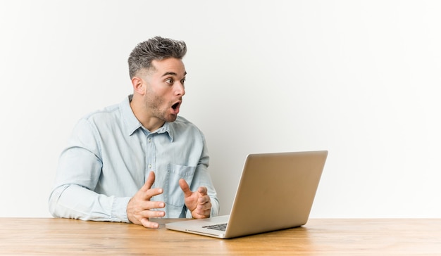 Junger hübscher Mann, der mit seinem Laptop arbeitet, der wegen etwas geschockt wird, das sie gesehen hat.