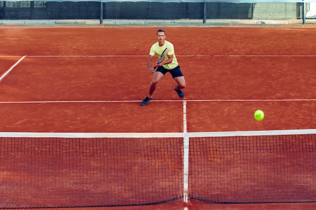 Junger hübscher Mann, der Tennis auf dem Sandtennisplatz spielt