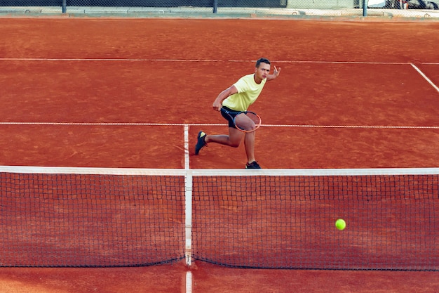 Junger hübscher Mann, der Tennis auf dem Sandtennisplatz spielt