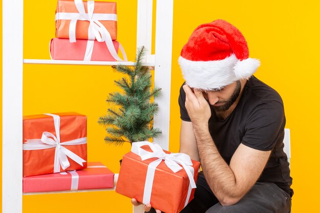 Junger hübscher Mann in einer Weihnachtsmannmütze hält Glückwunschgeschenke neben einem Weihnachtsbaum. Konzept der Feiertage von Weihnachten und Neujahr.