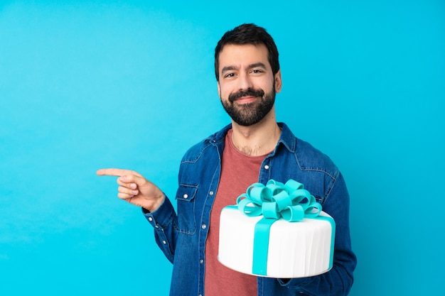 Junger hübscher Mann mit einem großen Kuchen über blau zeigendem Finger zur Seite