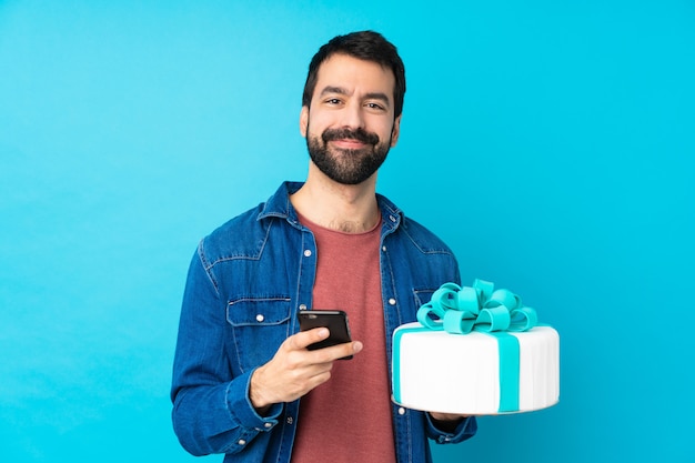 Junger hübscher Mann mit einem großen Kuchen über isolierter blauer Wand, die eine Nachricht mit dem Handy sendet