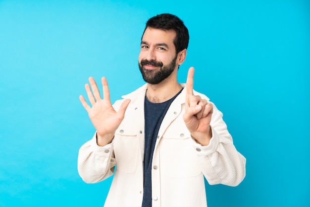 Junger hübscher Mann mit weißer Cordjacke über isolierter blauer Wand, die sechs mit den Fingern zählt