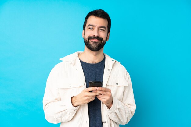 Junger hübscher Mann mit weißer Kordjacke über isolierter blauer Wand, die eine Nachricht mit dem Handy sendet