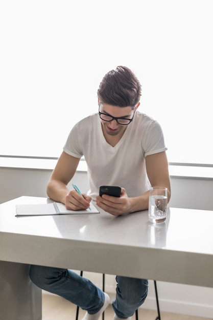 Junger hübscher Mann schreibt etwas auf Papiernotizbuch und hält mobiles Smartphone zu Hause. Unternehmenskonzept.