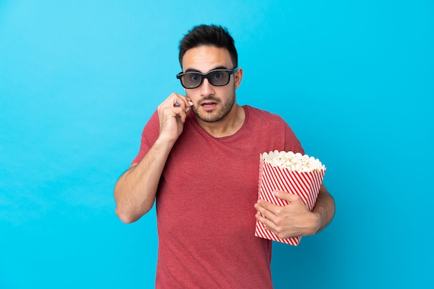 Junger hübscher Mann über Blau mit 3d Brille und hält einen großen Eimer Popcorn