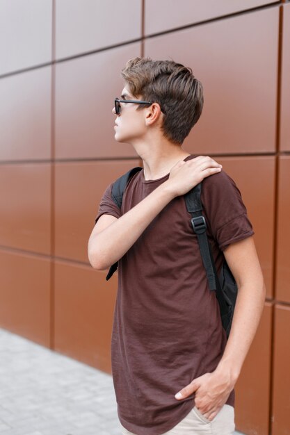 Junger hübscher moderner Hipster-Mann in der trendigen dunklen stilvollen Sonnenbrille in der trendigen Sommerkleidung mit einem schwarzen Rucksack geht an einem Sommertag durch die Stadt