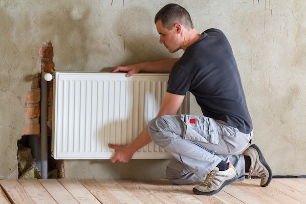 Junger hübscher professioneller Klempnerarbeiter, der Heizkörper in einem leeren Raum einer neu gebauten Wohnung oder eines neu gebauten Hauses installiert. Bau-, Wartungs- und Reparaturkonzept.