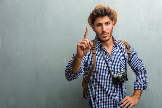 Junger hübscher Reisendmann, der einen Strohhut, einen Rucksack und eine Fotokamera zeigt, die num zeigen