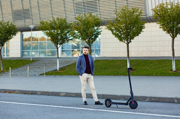 Junger hübscher stilvoller Mann mit E-Roller, der auf Bürgersteig nahe dem Flughafen auf dem Sonnenuntergang steht