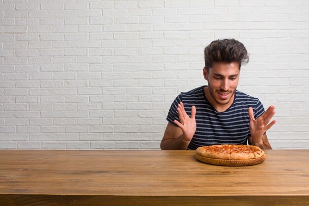Junger hübscher und natürlicher Mann, der auf einer Tabelle überrascht und entsetzt sitzt und mit großen Augen schaut, aufgeregt durch ein Angebot oder durch einen neuen Job, Gewinnkonzept. Eine leckere Pizza essen.