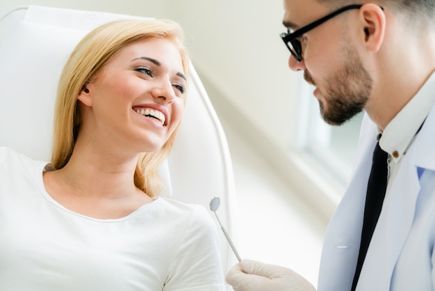 Junger hübscher Zahnarzt spricht mit dem glücklichen Frauenpatienten, der auf Zahnarztstuhl in der zahnmedizinischen Klinik sitzt.
