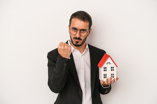 Junger Immobilienmaklermann, der ein Musterhaus lokalisiert auf weißem Hintergrund hält, das Faust zur Kamera, aggressiven Gesichtsausdruck zeigt.