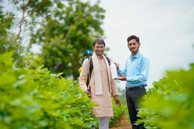 Junger indischer Agronom, der dem Landwirt eine Flüssigdüngerflasche gibt und Produktinformationen auf dem grünen Landwirtschaftsfeld sagt.