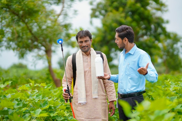 Junger indischer Agronom, der dem Landwirt eine Flüssigdüngerflasche gibt und Produktinformationen auf dem grünen Landwirtschaftsfeld sagt.