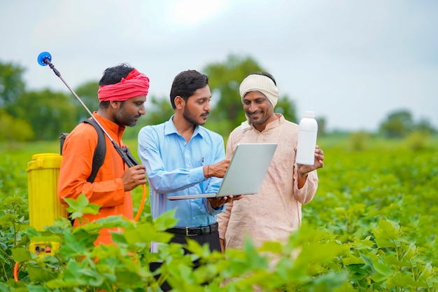 Junger indischer Agronom, der dem Landwirt eine Flüssigdüngerflasche gibt und Produktinformationen im Laptop auf dem grünen Landwirtschaftsfeld zeigt.