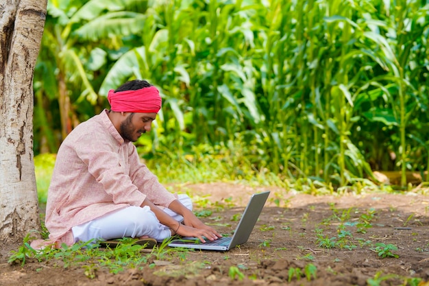 Junger indischer Bauer, der Laptop am Landwirtschaftsfeld verwendet.