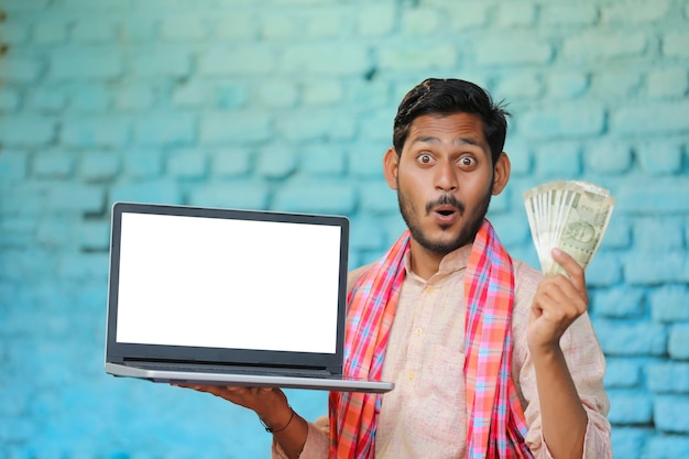Junger indischer Bauer, der Laptopbildschirm und Geld zeigt