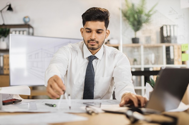 Junger indischer Bauingenieur in weißem Hemd mit Krawatte sitzt auf dem Schreibtisch im Büro