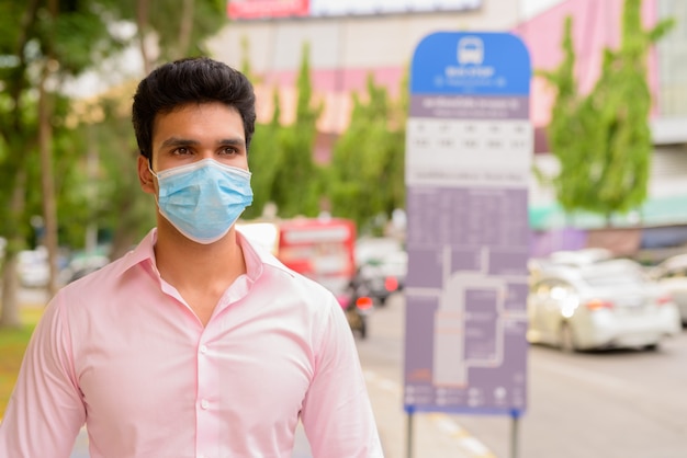 Junger indischer Geschäftsmann, der Maske trägt, während an der Bushaltestelle wartet
