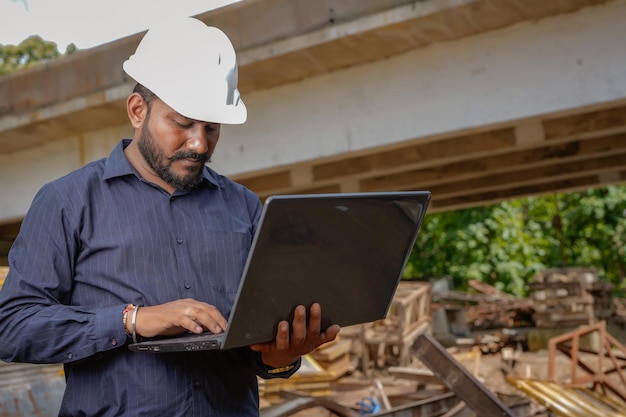 Junger indischer Ingenieur in Uniform und mit Laptop im Feld