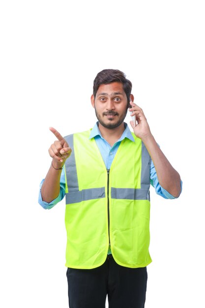 Junger indischer Ingenieur mit Hardhat und sprechendem Smartphone auf weißem Hintergrund.