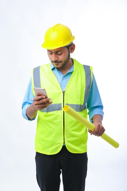 Junger indischer Ingenieur mit Helm und Smartphone auf weißem Hintergrund.