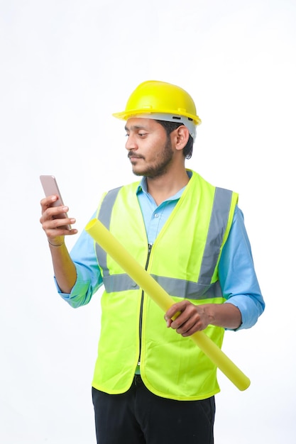 Junger indischer Ingenieur mit Helm und Smartphone auf weißem Hintergrund.