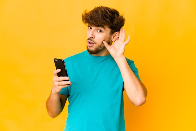 Junger indischer Mann, der ein Telefon hält, das versucht, einen Klatsch zu hören.